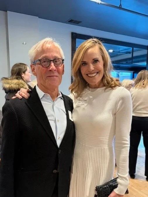A smiling young woman in a white dress with blonde hair is standing next to an older man in glasses, a white shirt, and a black jacket, with her arm around him, at an indoor event with other people in the background.