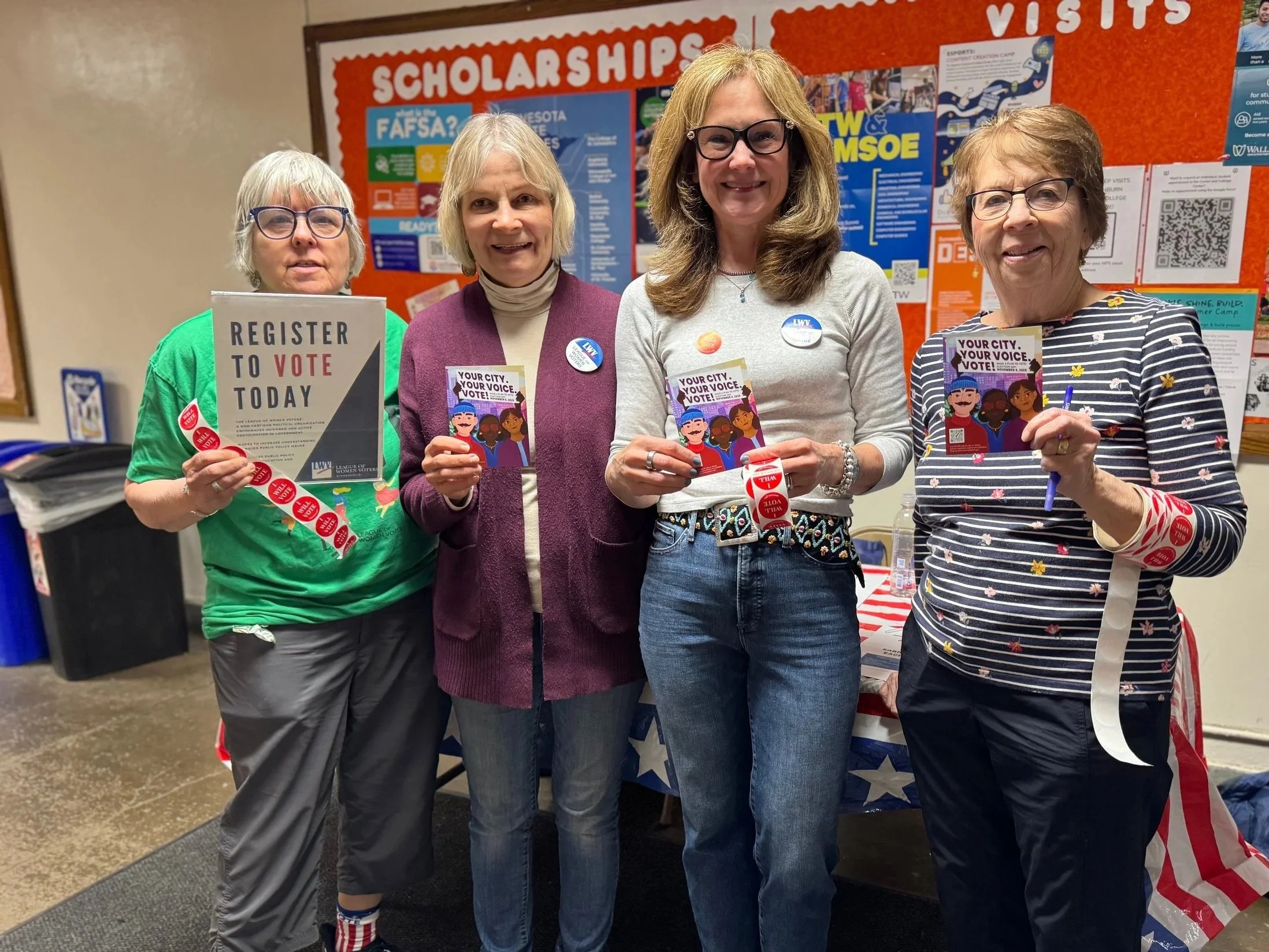 Four women standing indoors, holding flyers about voting and scholarships, smiling at the camera. Behind them is a bulletin board with information about scholarships and visits.