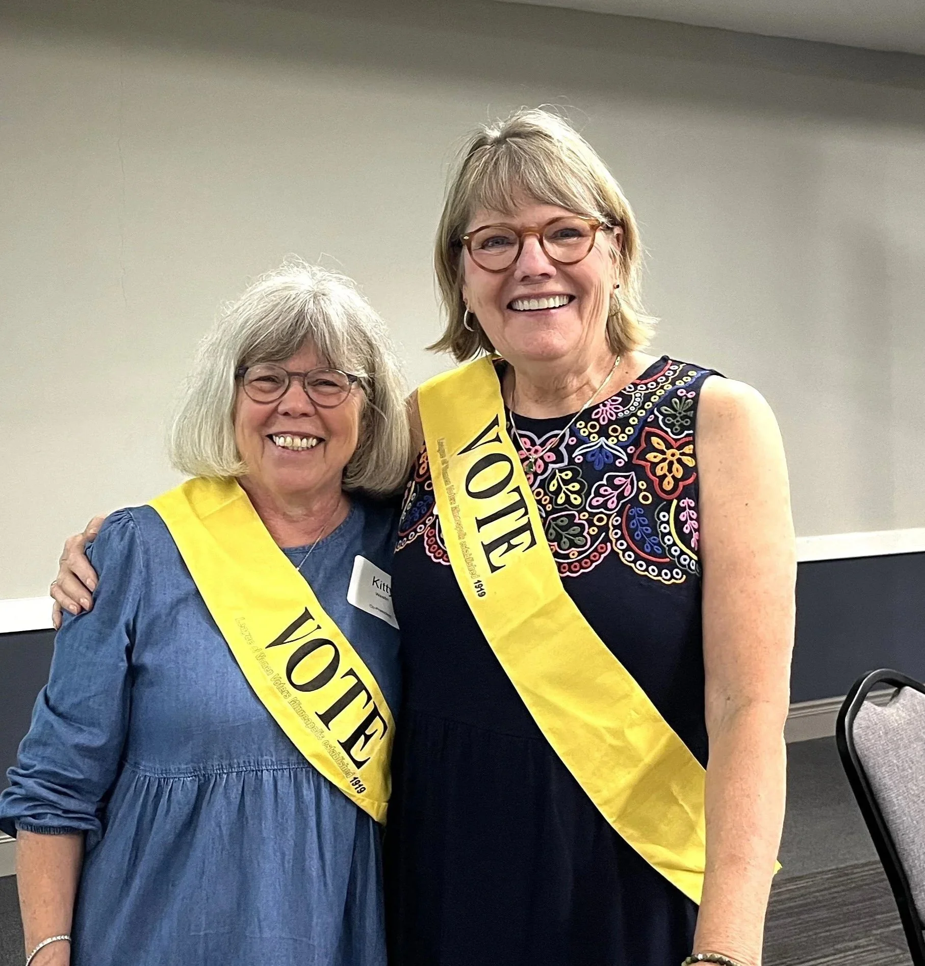 Two women smiling with yellow sashes, one wearing glasses and a blue top and the other wearing glasses and a colorful embroidered dress, posing together indoors.