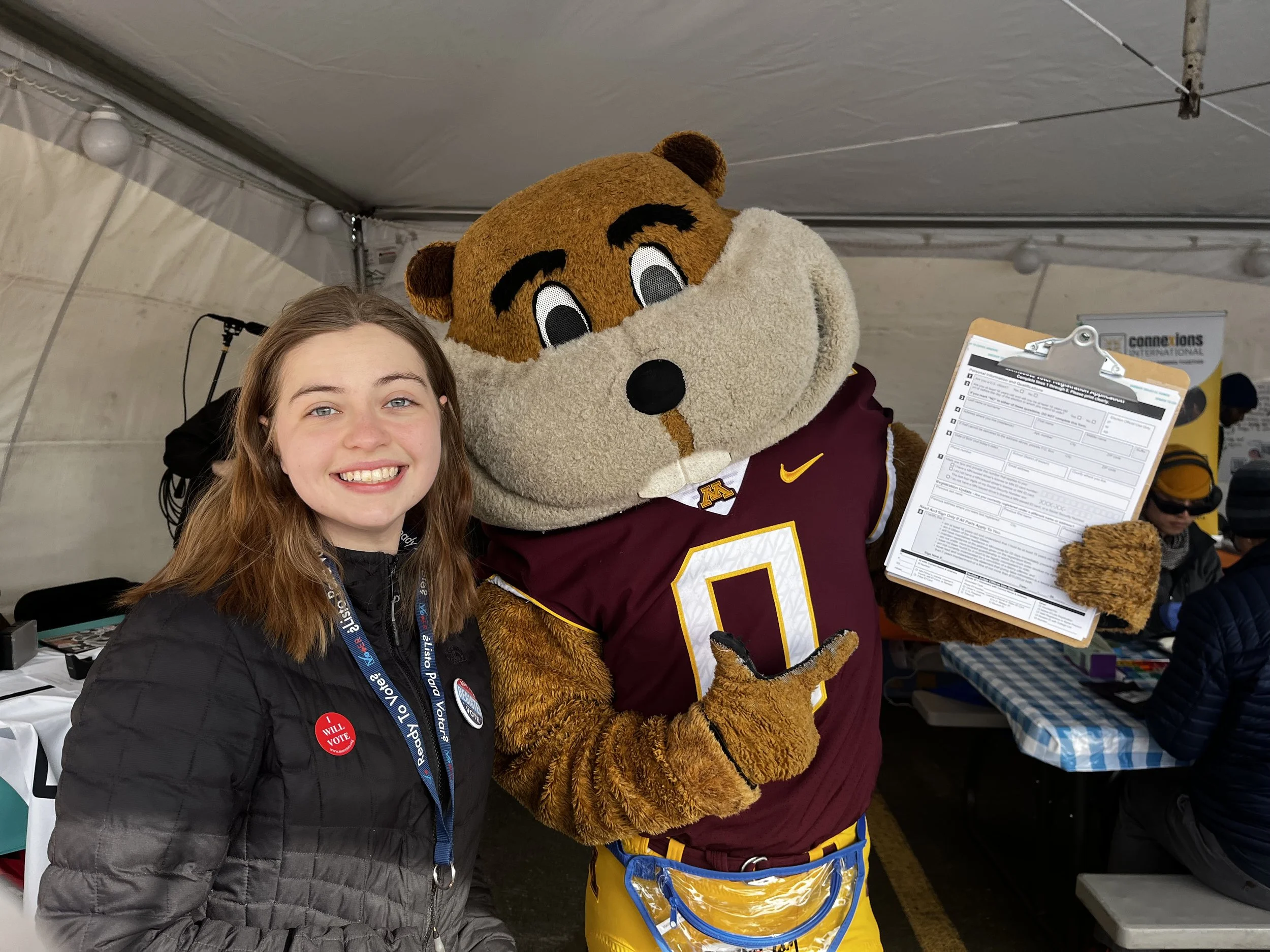 A young woman with long brown hair smiling at the camera standing next to a person in a mascot costume of a bear, who is holding a clipboard. The mascot is wearing cardinal and gold sportswear, representing the University of Minnesota. The scene appears to be at an indoor event or gathering with tables and other people in the background.