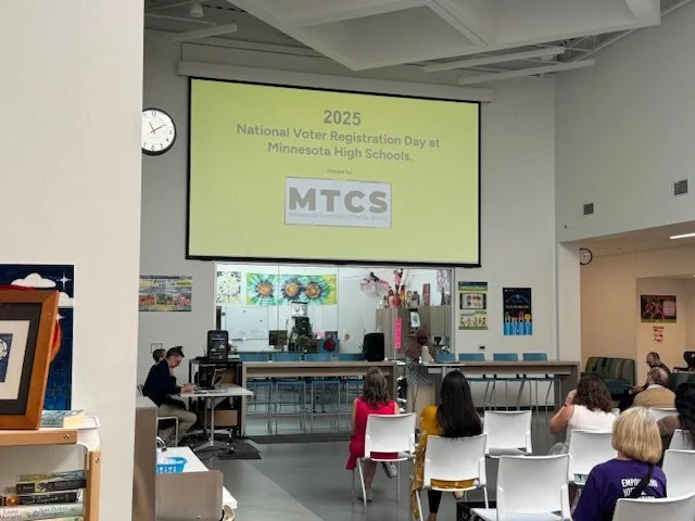 A classroom or auditorium with people seated facing a large yellow screen that displays '2025 National Voter Registration Day at Minnesota High Schools.' There are decorations and flowers in front of the screen, and the room has a clock on the wall s
