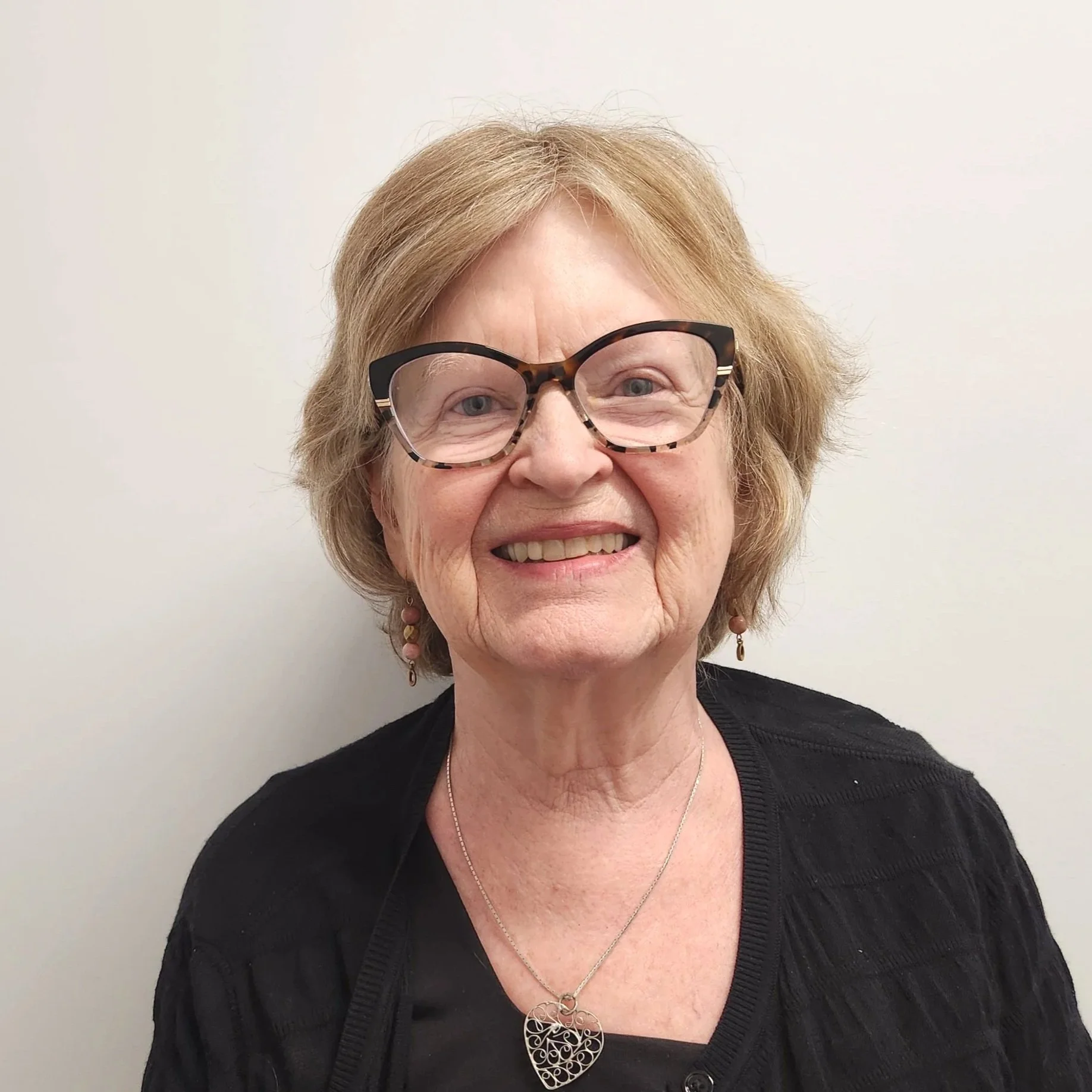 A woman with curly, reddish-blonde hair and glasses with black and tortoise shell frames, smiling and showing her teeth, against a plain white wall. She is wearing a black top, a silver necklace with a heart-shaped pendant, and dangling earrings.