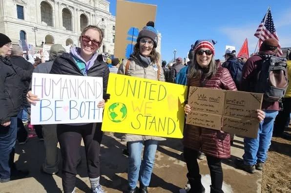Three women holding signs at a rally, with national monuments and other protesters in the background.