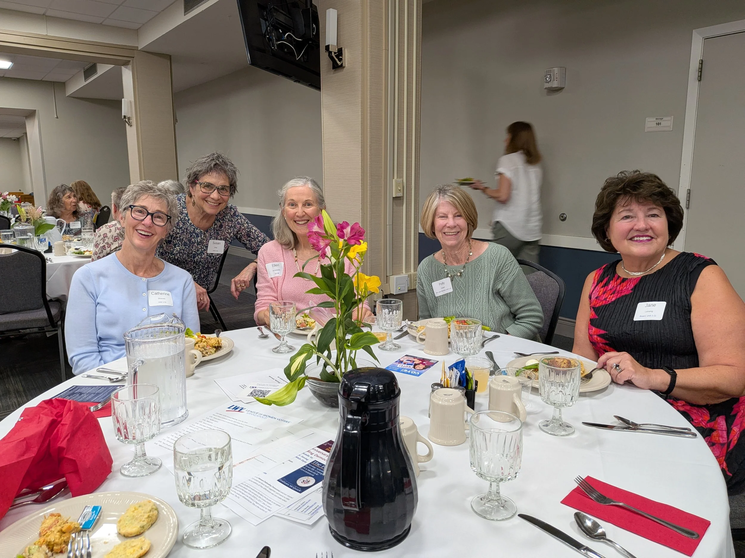 Women gathered around a dining table at a formal event, smiling, with plates of food, drinks, and flowers on the table.