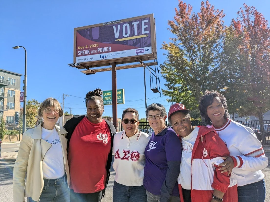 Six diverse women standing together outdoors on a sunny day, smiling with trees and a billboard that says 'VOTE' in the background.