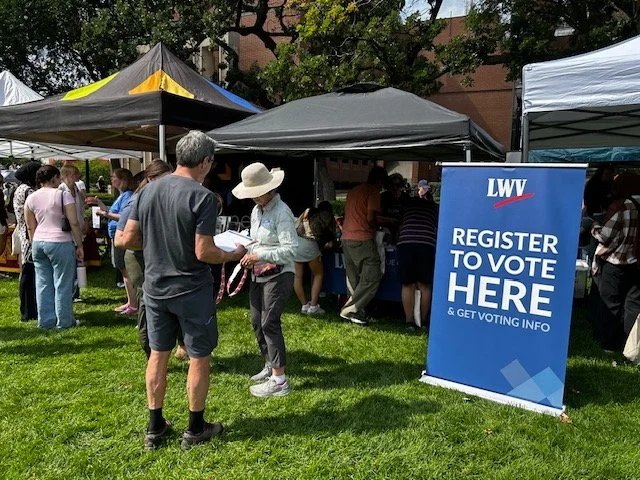 People gathered at an outdoor voting registration booth, with tents and a large blue sign displaying 'LWV REGISTER TO VOTE HERE & GET VOTING INFO'.