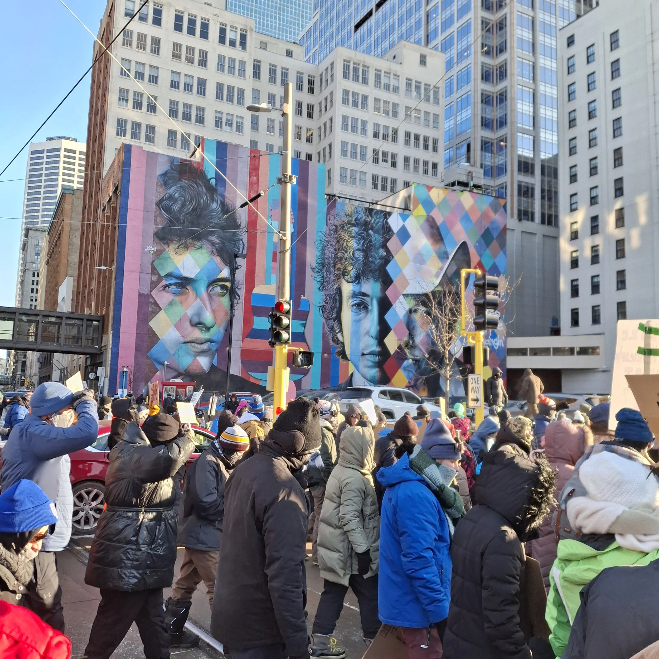 Crowd of people wearing winter clothing gathered on a city street with tall buildings and a large colorful mural depicting faces in the background.