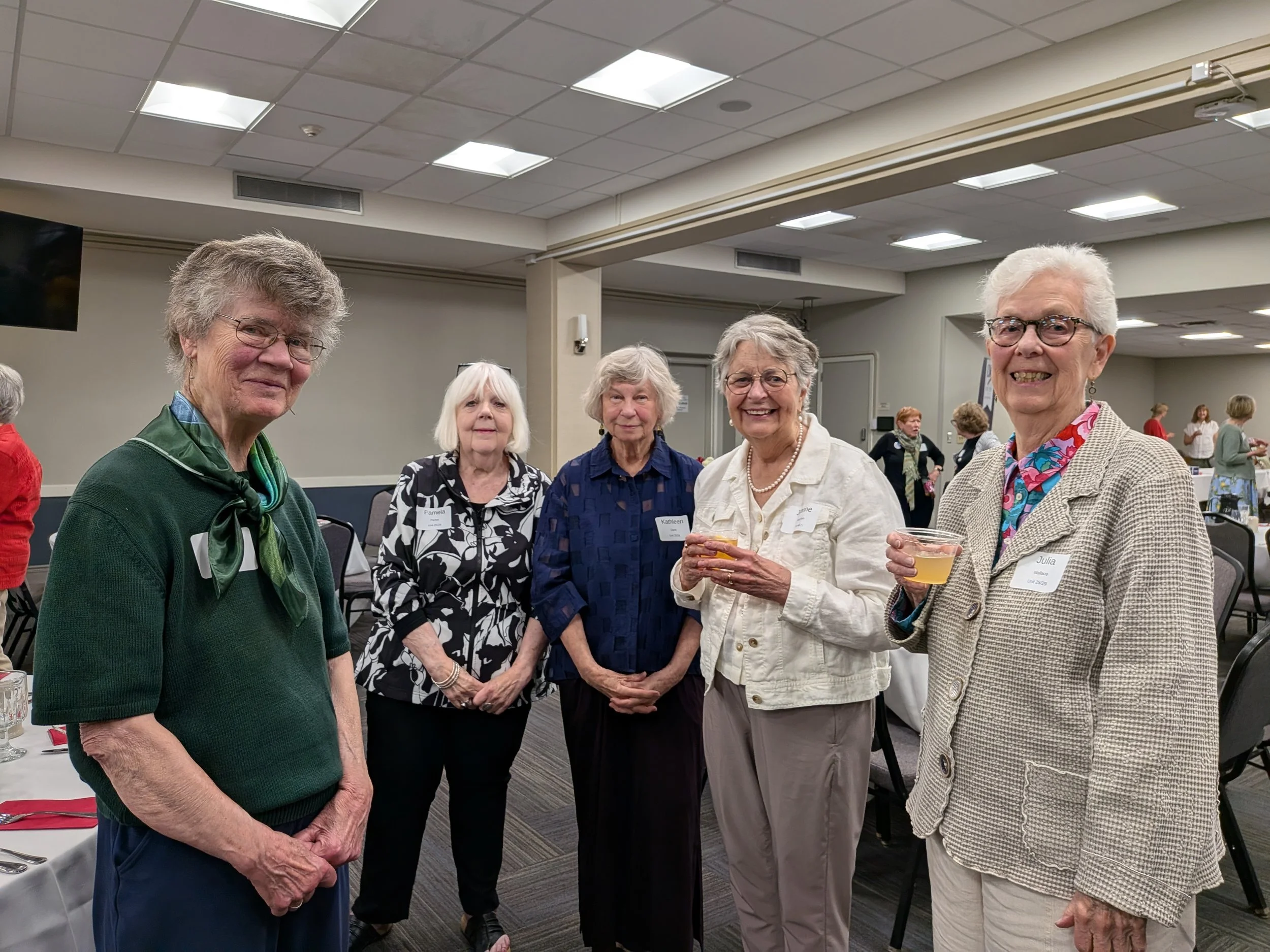 Group of six elderly women standing together in a banquet hall, smiling, some holding drinks, dressed in semi-formal attire, with other guests in the background.