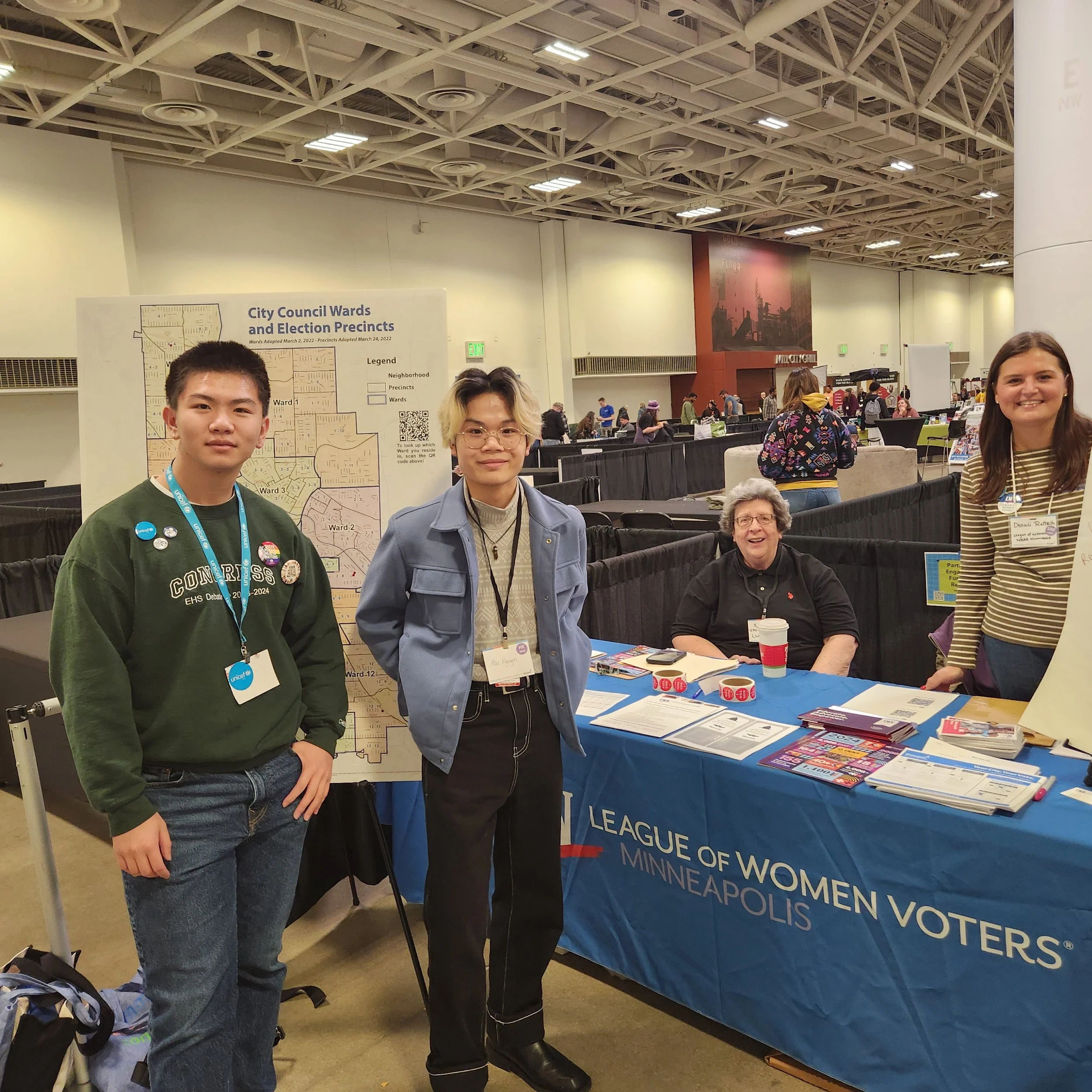 People standing at a booth for the League of Women Voters at an indoor event, with a large poster displaying a map and information about City Council Wards and Election Precincts in Minneapolis, Minnesota in the background.
