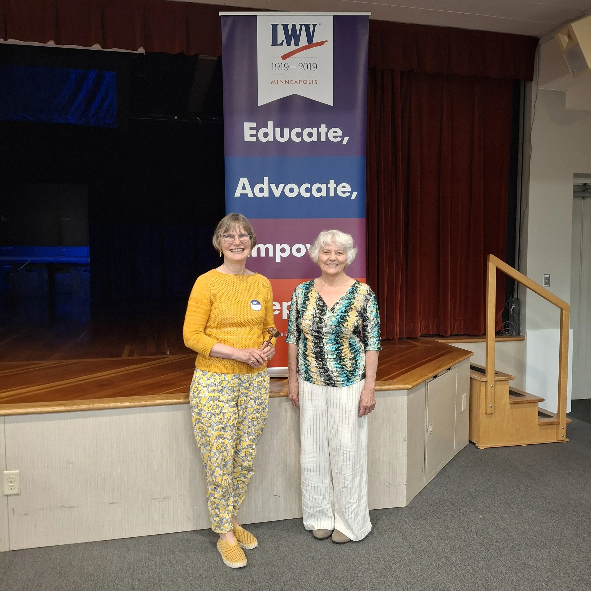 Two women standing in front of a purple banner with the LWV logo and the words 'Educate, Advocate,'. One woman is wearing a yellow sweater and patterned yellow pants, while the other is dressed in a floral patterned blouse and white pants. They are s