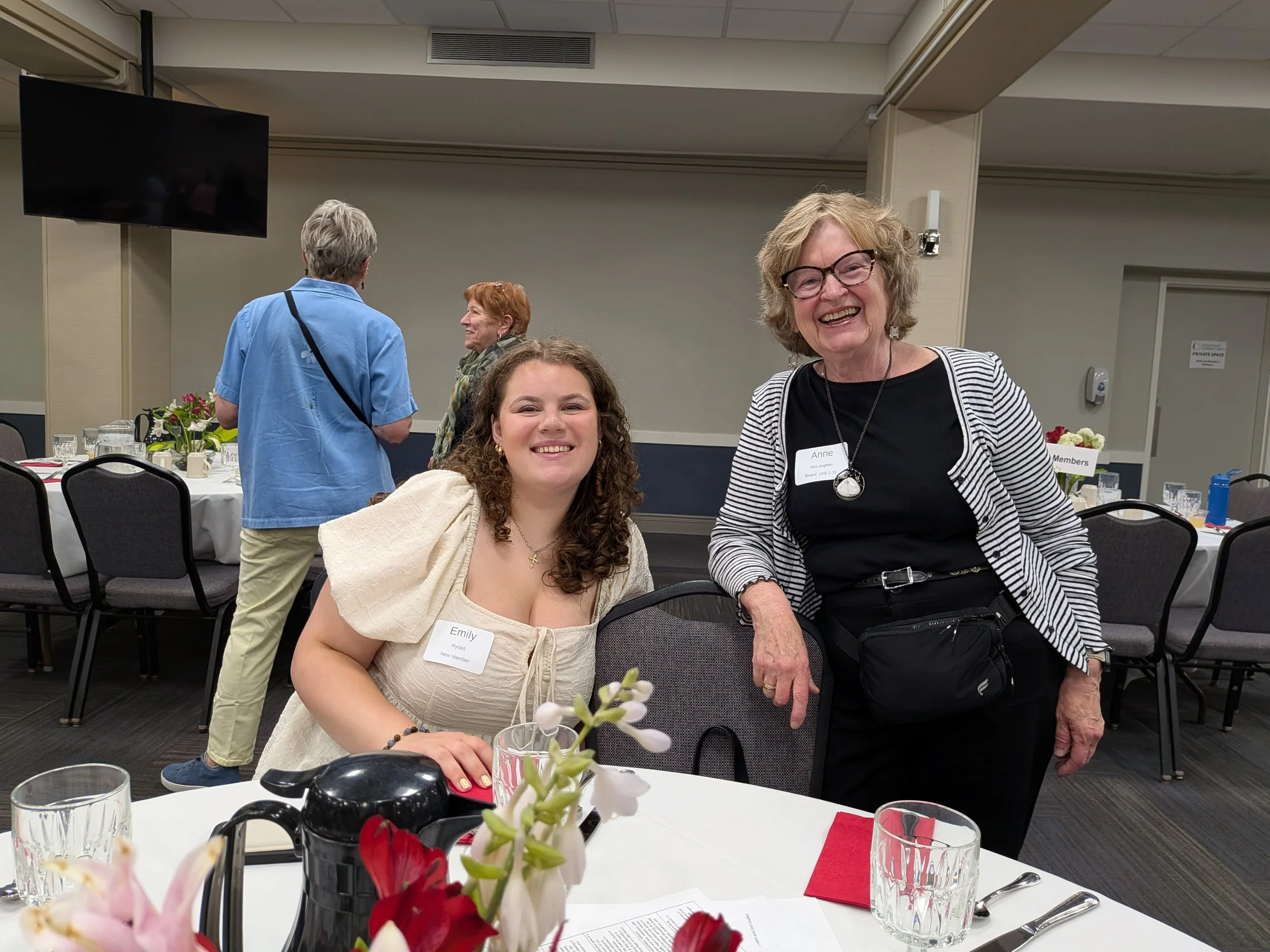 A group of women at a formal gathering in a banquet hall, smiling and interacting. Two women are in the foreground, one with curly hair and a cream-colored dress, and the other with glasses and a striped blazer. Other women are in the background, tal