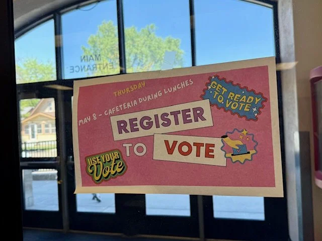 A colorful sign inside a building, encouraging people to register to vote during lunch on Thursday, May 8, with phrases like "Get Ready to Vote" and "Use Your Vote," and a graphic of a hand casting a ballot.