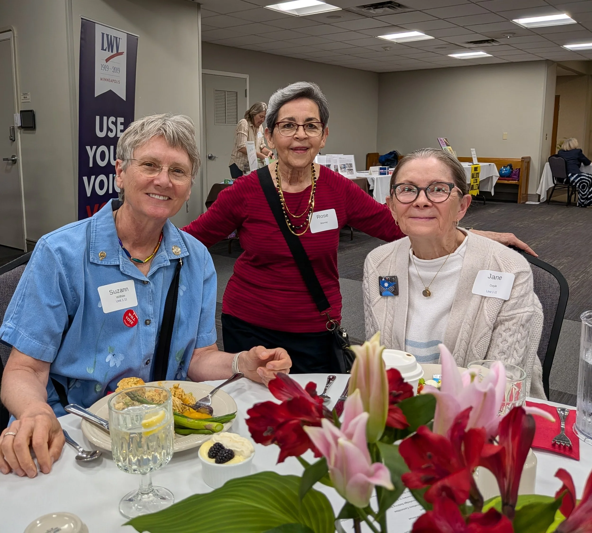 Three women are smiling at a round table decorated with a pink and red flower arrangement in a banquet hall. The woman in the middle wears a red top and glasses, while the women on either side wear light-colored clothing and name tags. There are plat