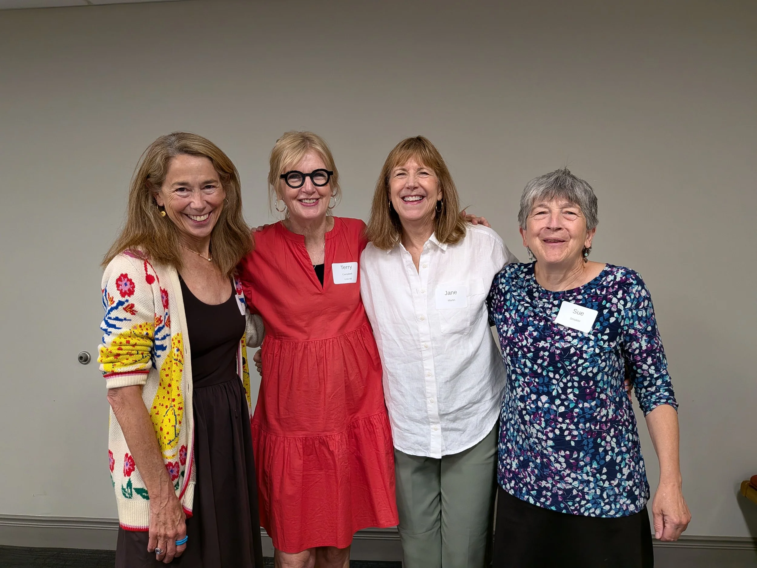 Four women standing together, smiling, with two wearing glasses and having name tags. They are dressed casually and standing against a plain gray wall.