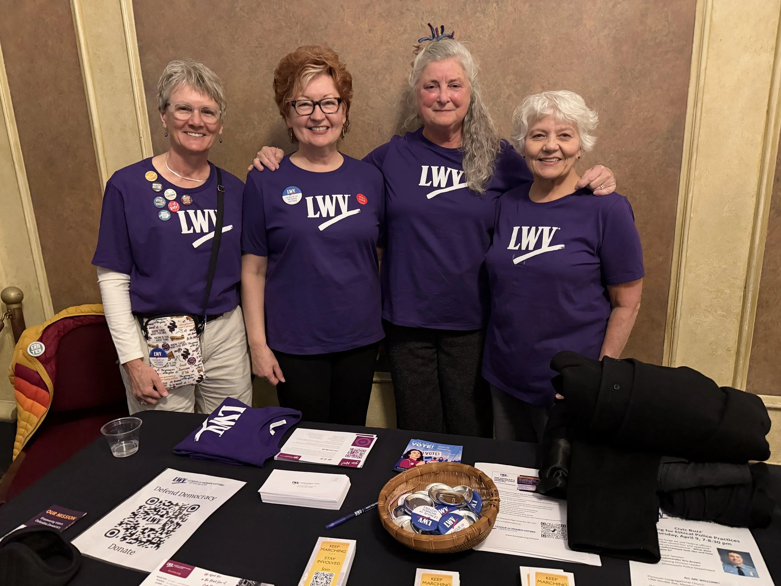 Four women standing together at a table for a voting or political event, wearing purple shirts with white text reading 'LWV', surrounded by campaign materials, buttons, flyers, and QR codes.