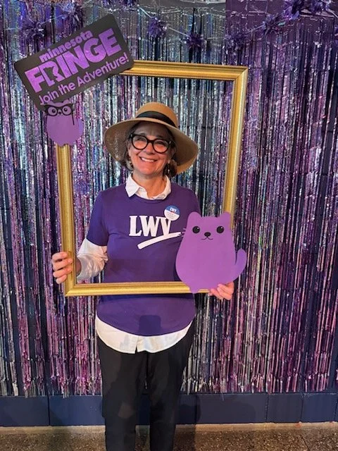 Woman with glasses and a hat holding a large photo frame, standing in front of a colorful, glittery backdrop. She is wearing a purple t-shirt with white letters, and is holding a purple cartoon cat cutout. There are signs that read "Minnesota Flinge" and "Join the Adventure!" in the background.