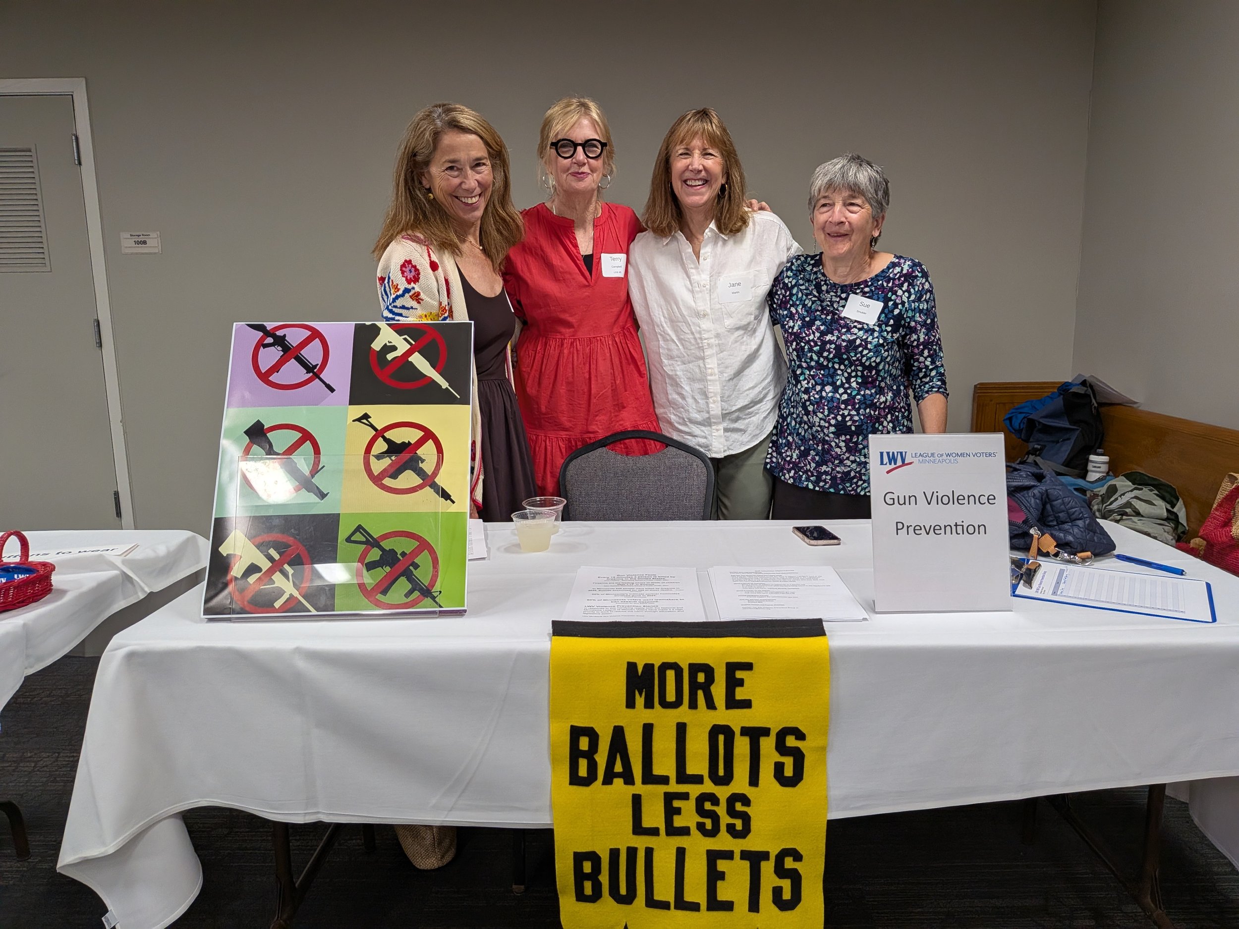 Four women standing behind a table with a sign that reads 'More Ballots Less Bullets' and display with illustrations of guns and crossed-out symbols, at a gun violence prevention event. The women are smiling and wearing name tags.