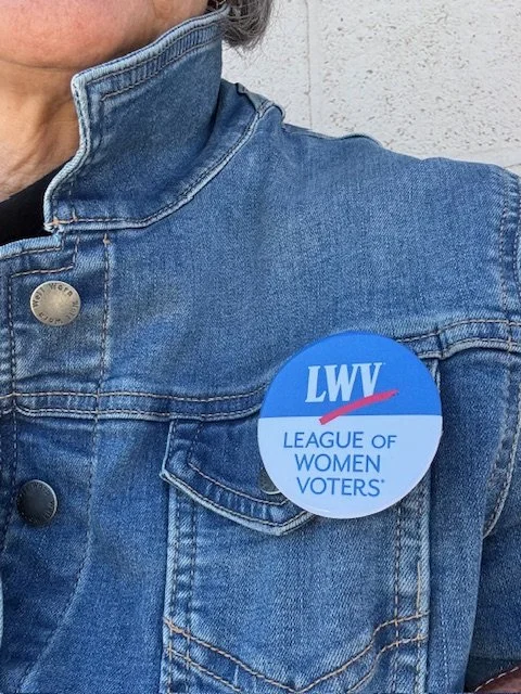 Close-up of a person wearing a denim jacket with a blue 'League of Women Voters' campaign button on the chest.