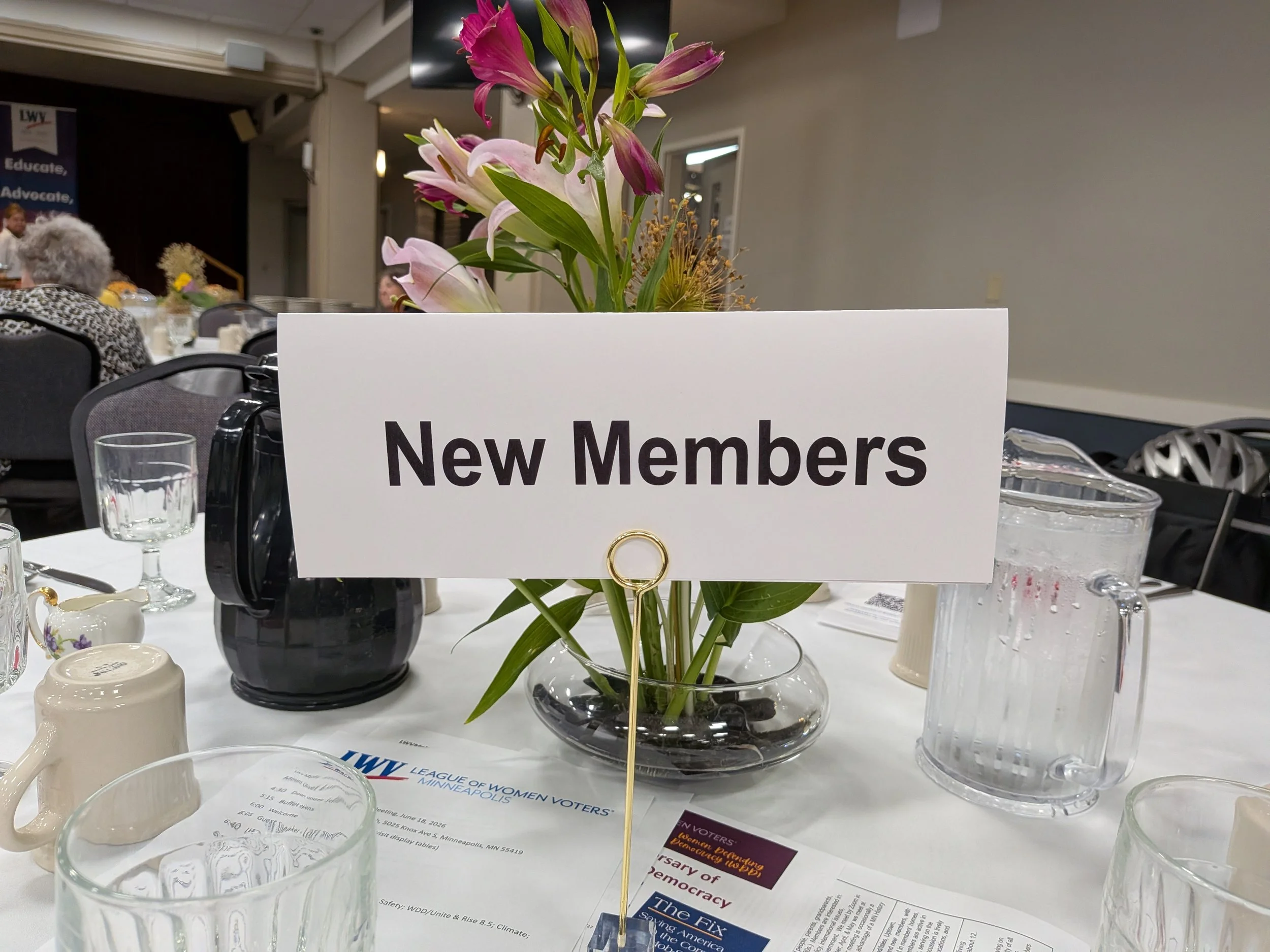 A glass vase with pink and white lilies and yellow accents on a table at a conference. There are cups, papers, and water glasses on the table, and a sign reading 'New Members' in front of the vase. In the background, people are seated at other tables