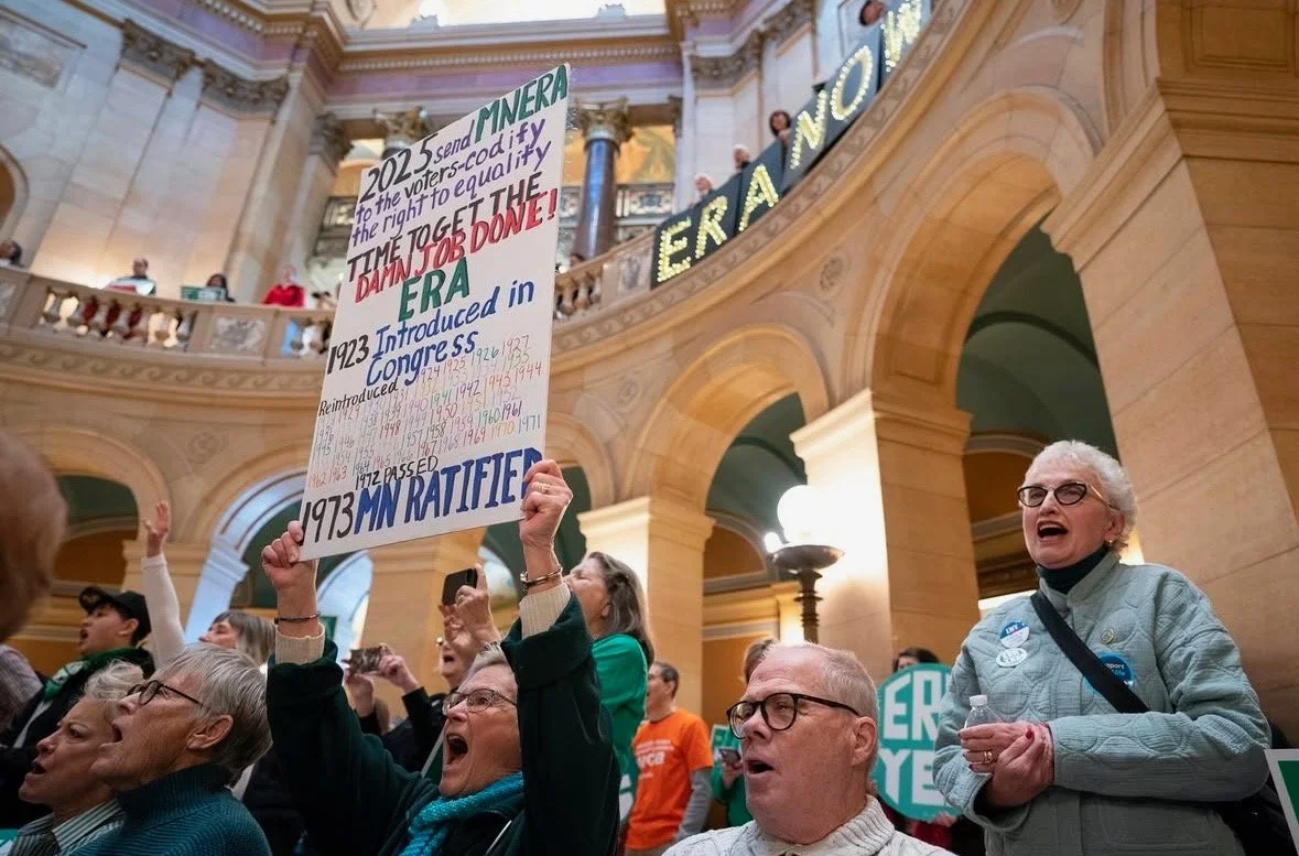 A group of people at a political rally inside a large building with arches, holding signs and protesting for voting rights and equal rights, with some individuals cheering and shouting.