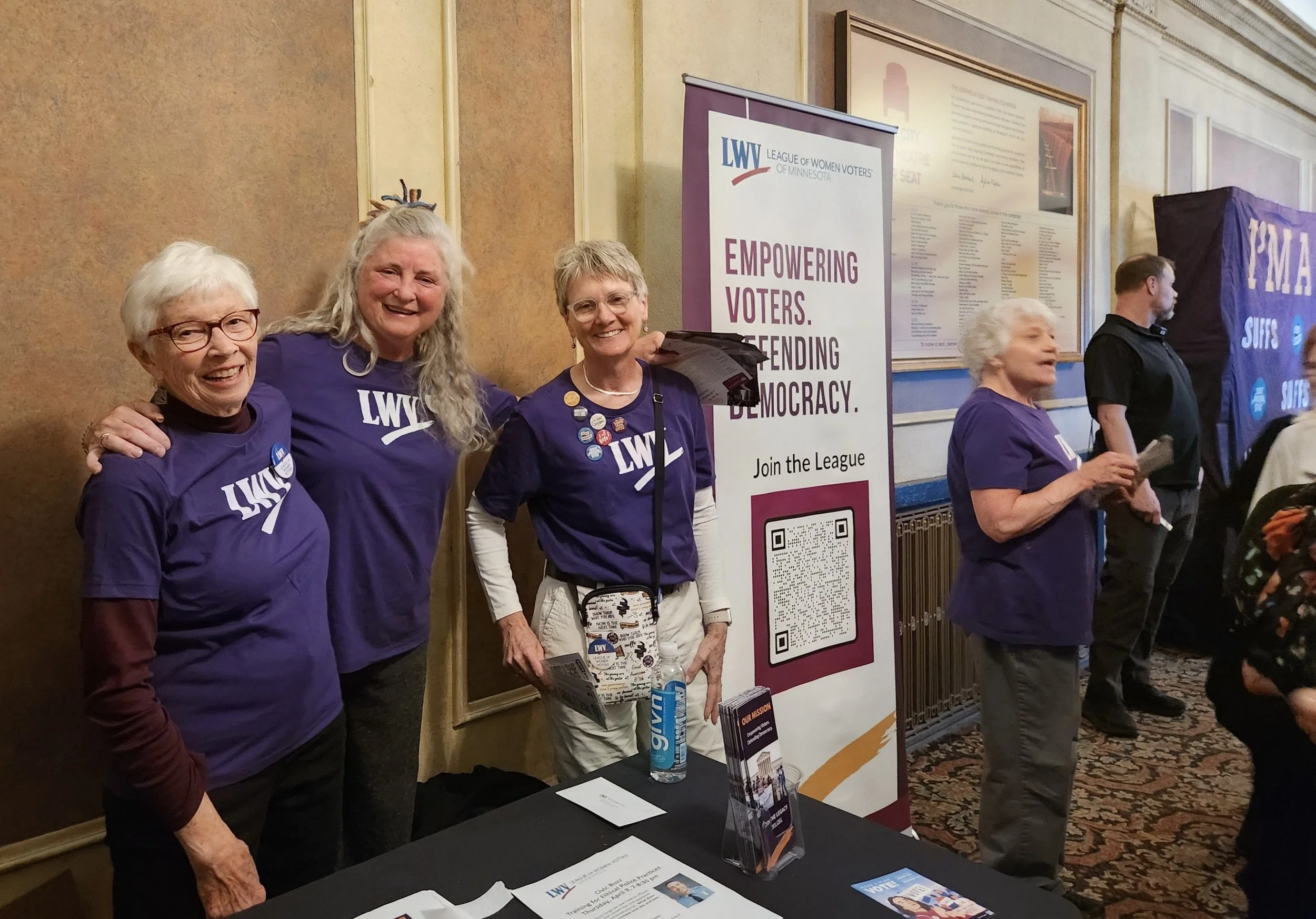 A group of people at a voter engagement event, wearing purple shirts with 'LWV' on them, standing next to a tall banner that reads 'Empowering Voters. Defending Democracy.' There are informational materials on the table and behind the group, includin