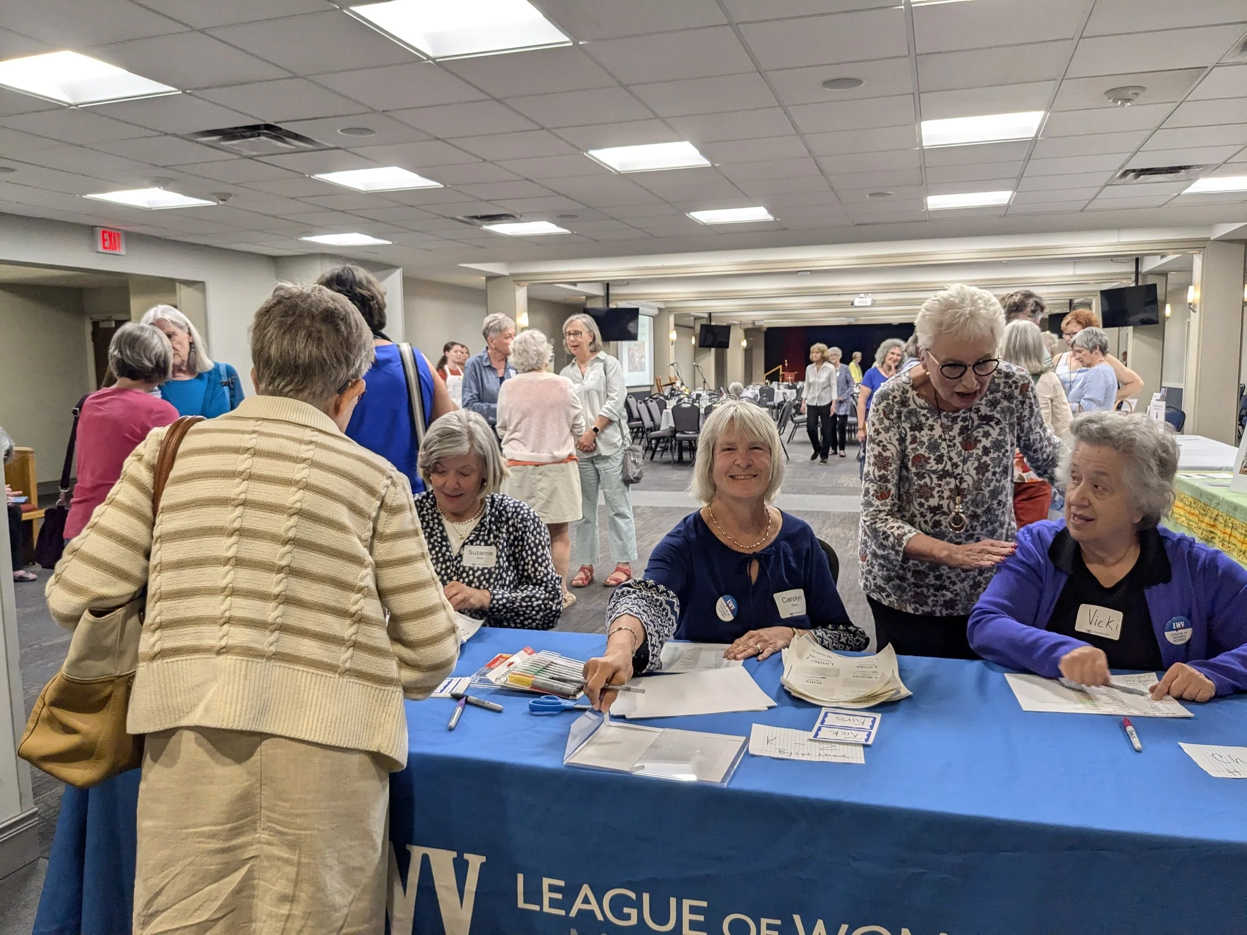 People gathered around a table with registration materials at a conference or event, with chairs and screens in the background.