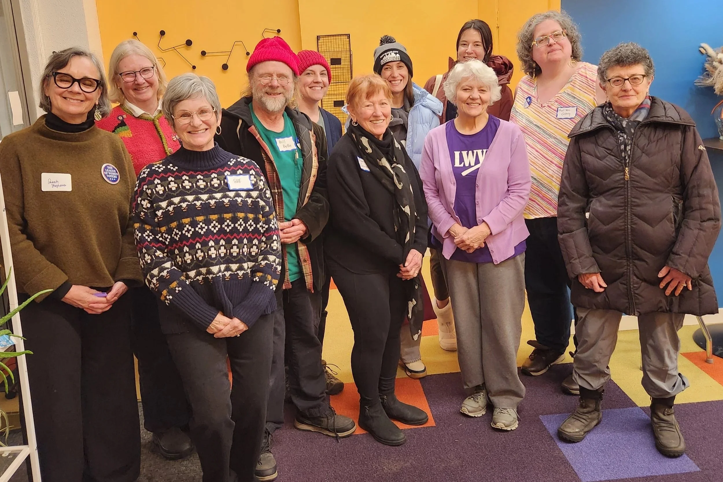 A group of fourteen diverse women standing together indoors against a colorful background, smiling at the camera.