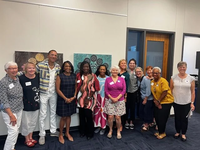 Group of fourteen diverse women standing together indoors, smiling, with artwork on the wall behind them.