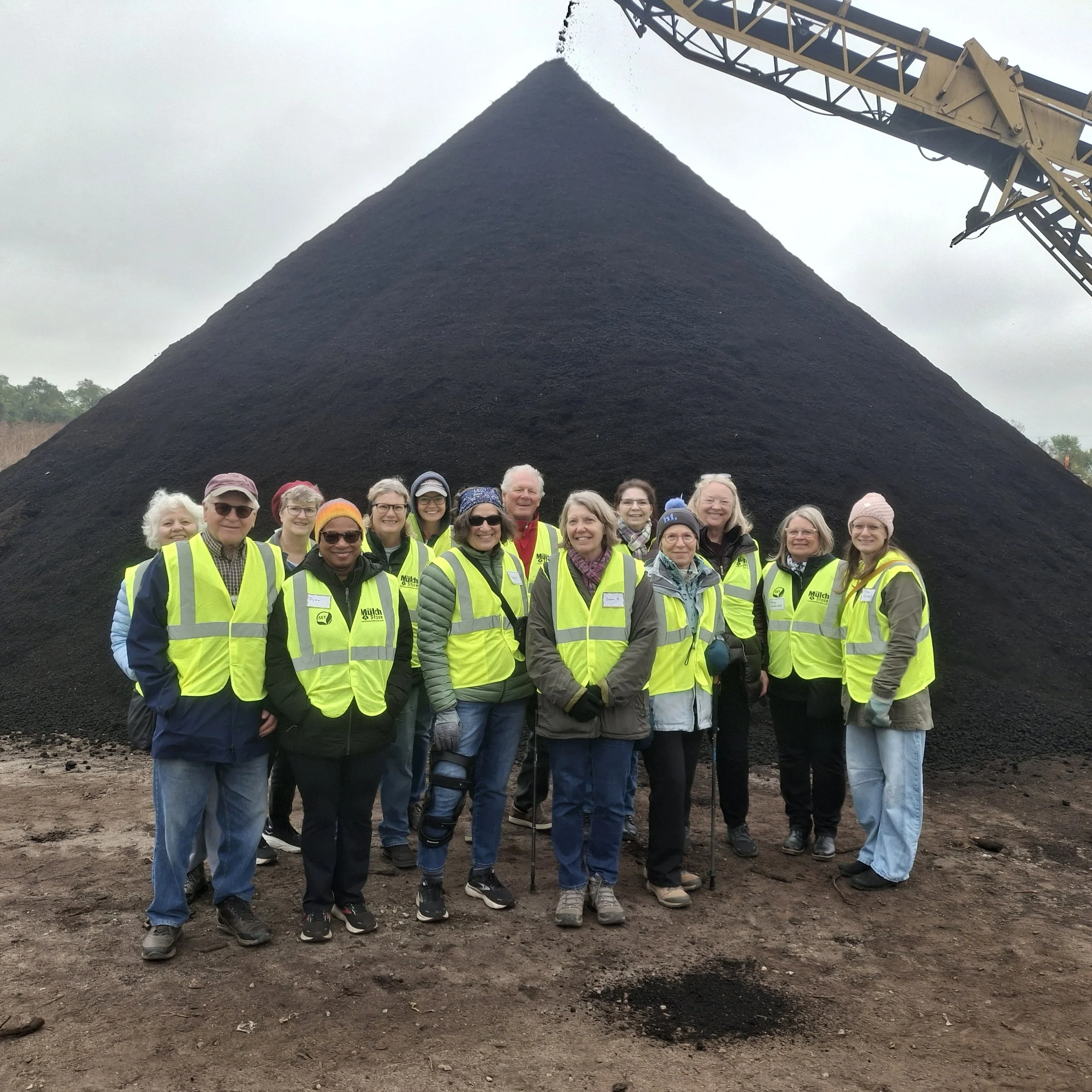 Group of people wearing yellow safety vests posing in front of a large pile of black coal with a conveyor belt above it.