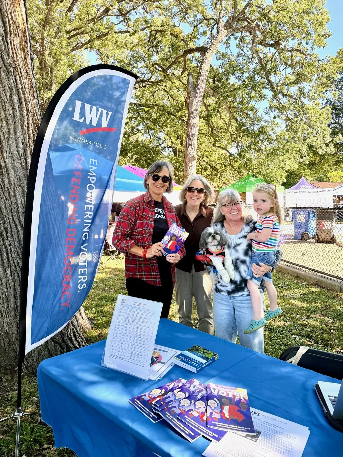 Four women and one young girl at a voting awareness event outdoors, with a flag and informational materials on a table, surrounded by trees and tents.