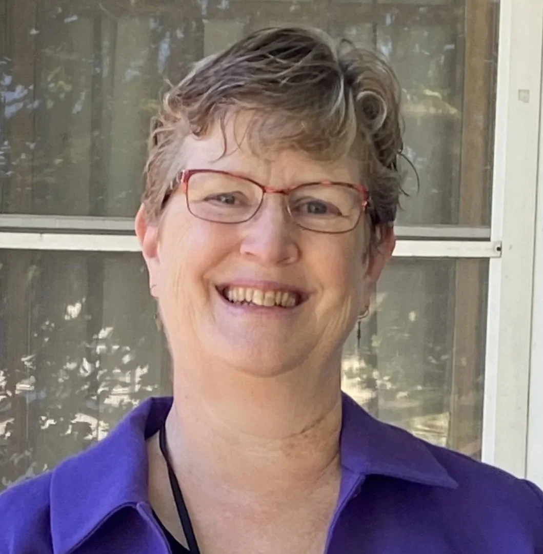 A woman with short, curly hair wearing glasses and a purple shirt, smiling outside in front of a window with trees reflected in it.