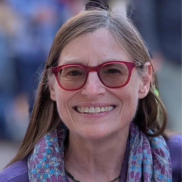 A woman with blonde hair, wearing red glasses, smiling, dressed in a purple top and a patterned scarf.