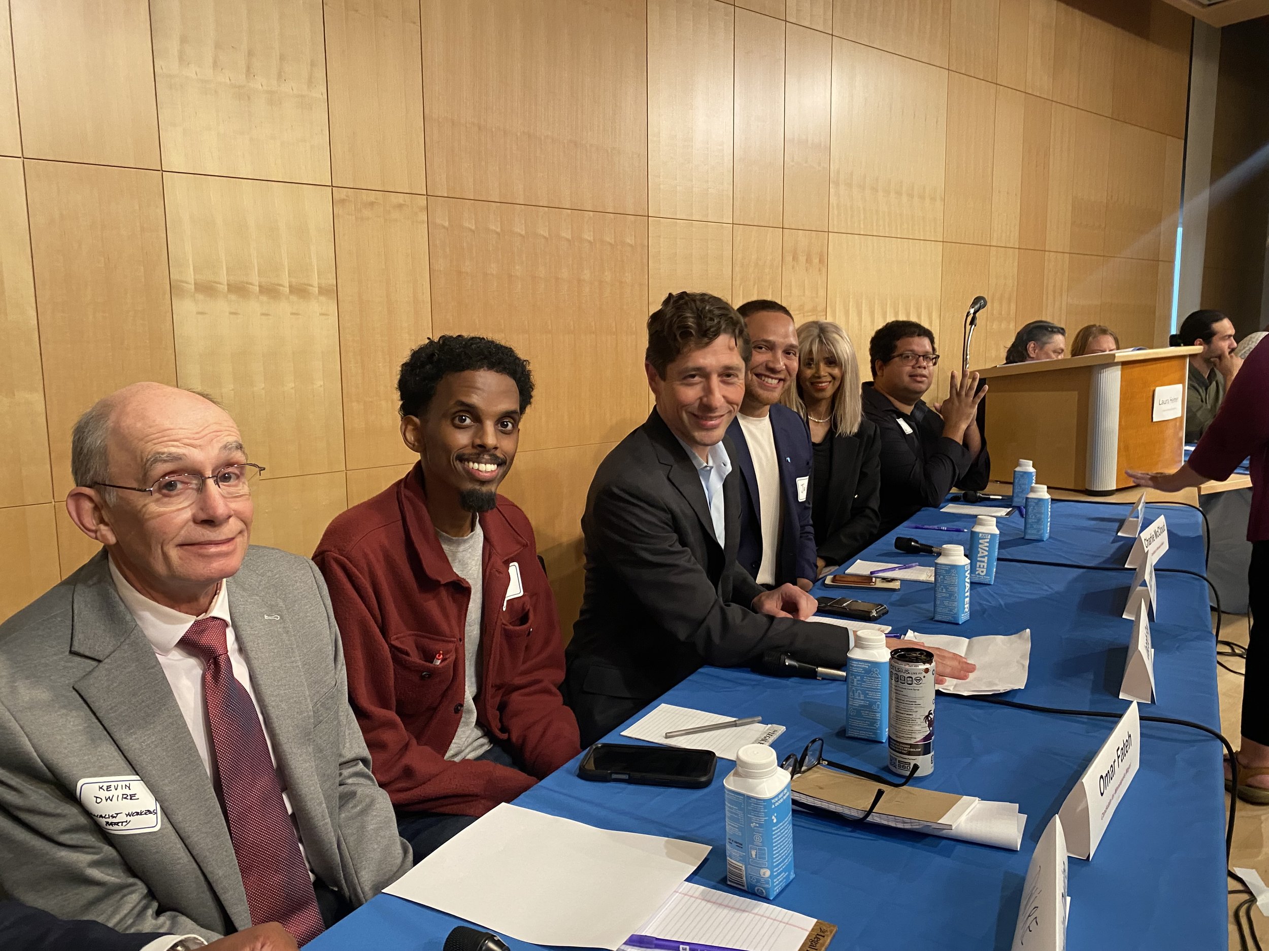 A group of diverse people sitting at a long table during a panel discussion or conference. There are water bottles, notepads, and name cards on the table. The background features a wooden wall.
