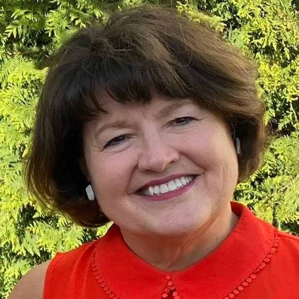 Smiling woman with short brown hair, wearing a red sleeveless top, standing outdoors with green foliage in the background.