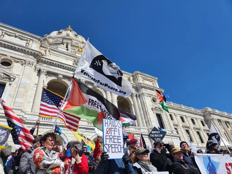 Protesters gather in front of a large government building, holding flags and signs supporting free speech, including a sign that reads ''Hands Off, Free Speech.'' Flags include the American flag and others with various symbols and colors.