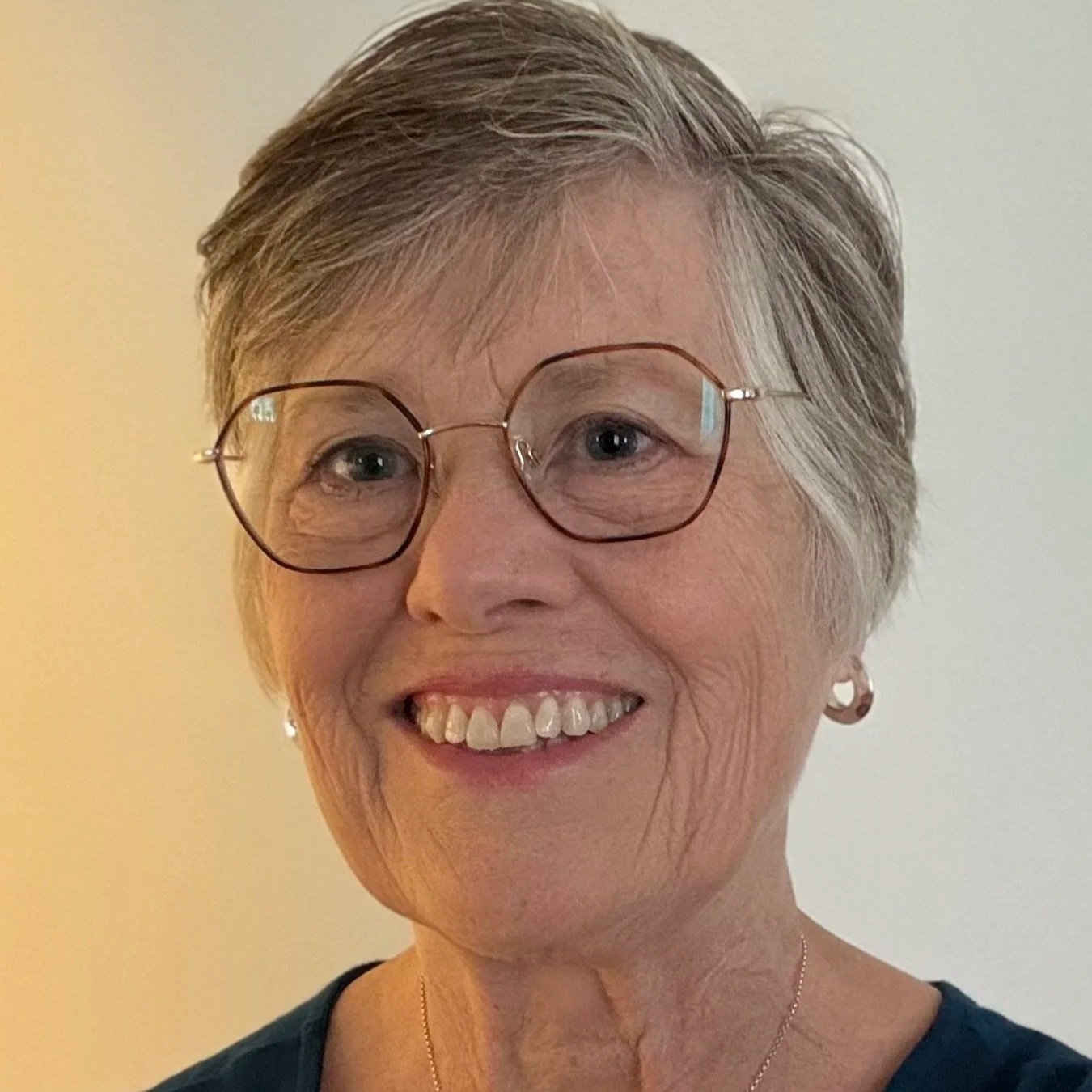 A smiling older woman with short gray hair, wearing glasses, earrings, a necklace, and a dark top, standing against a plain background.