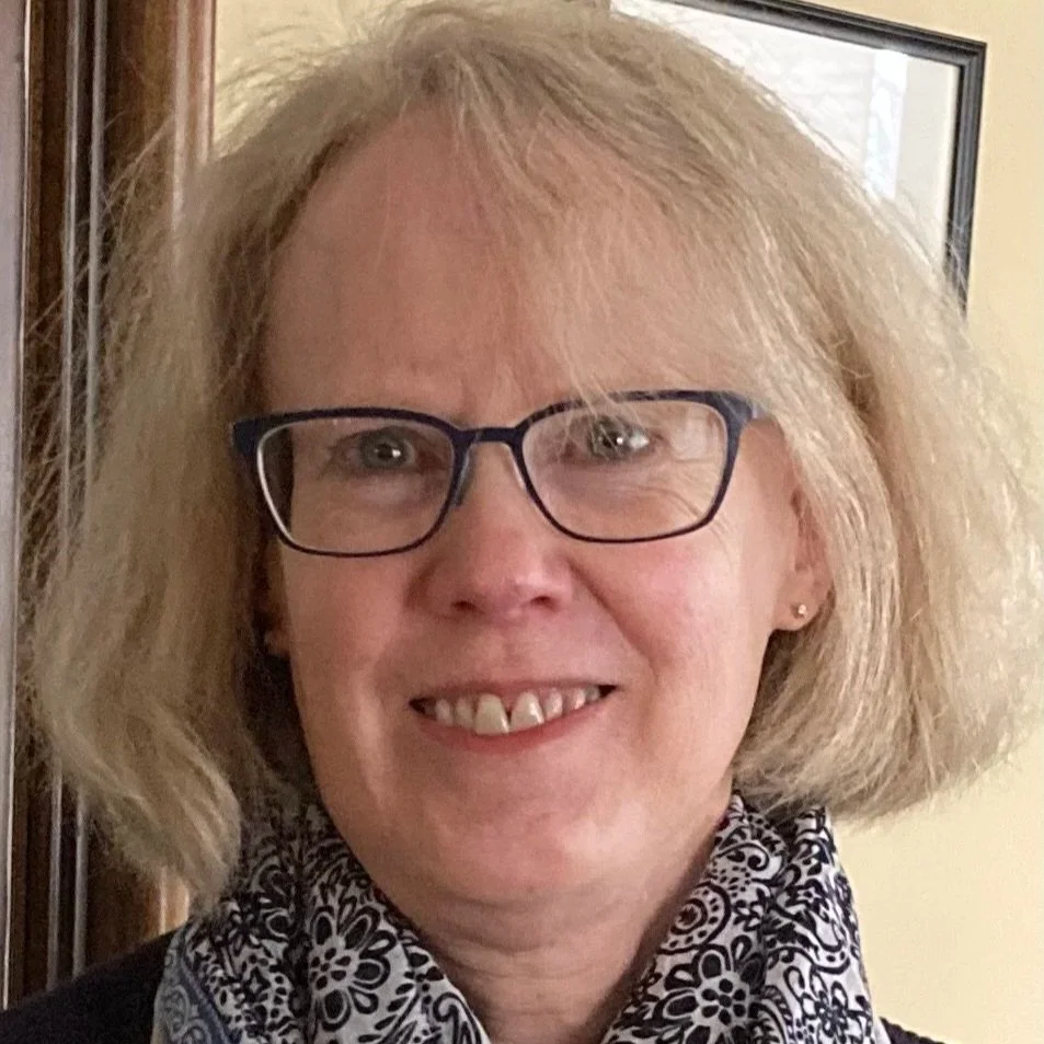 A woman with blonde hair, glasses, and a patterned scarf smiling indoors near a framed picture.