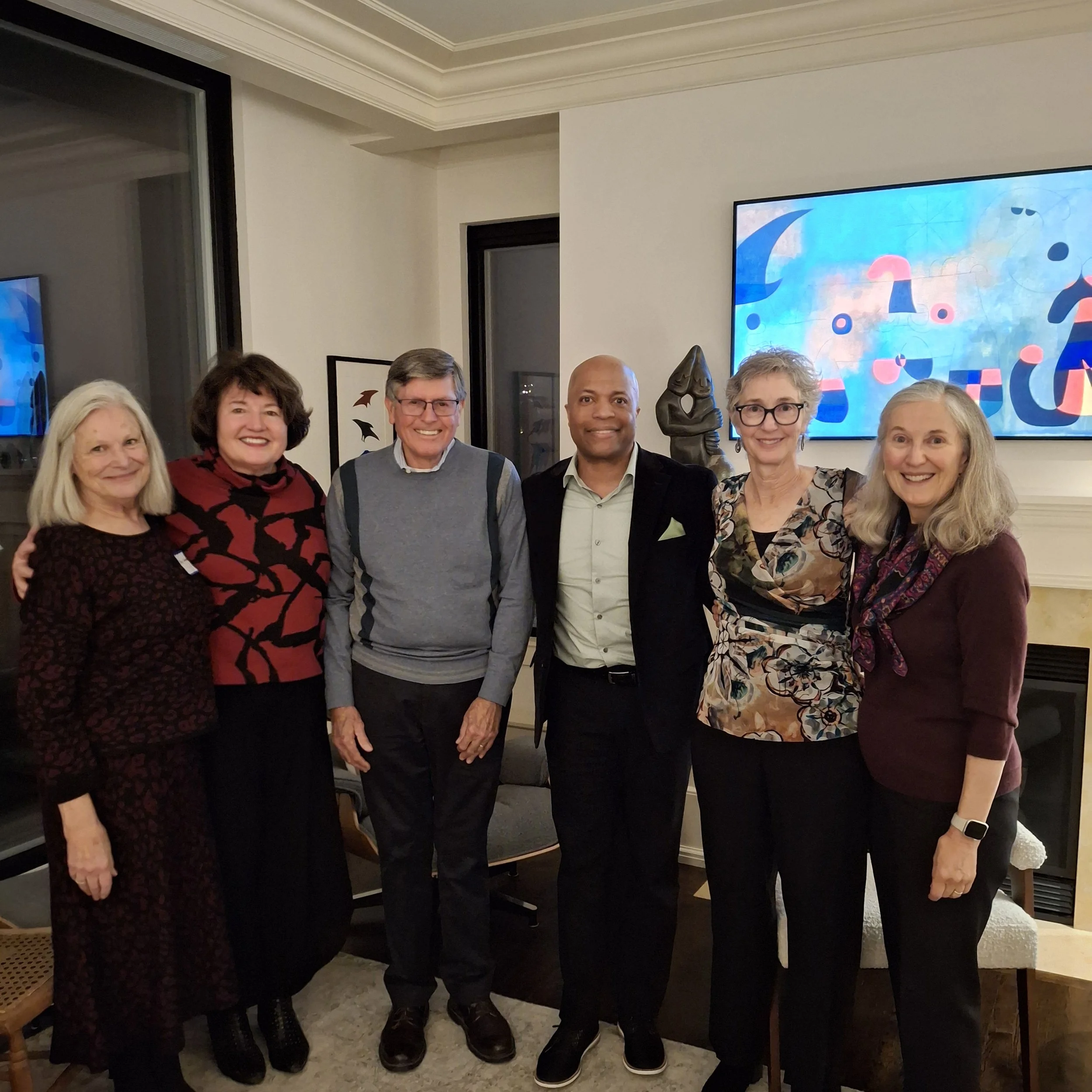 A group of seven smiling people standing closely together indoors in front of a wall with a large colorful abstract painting and a television screen. The group includes four women and three men, all dressed in casual to business casual attire.