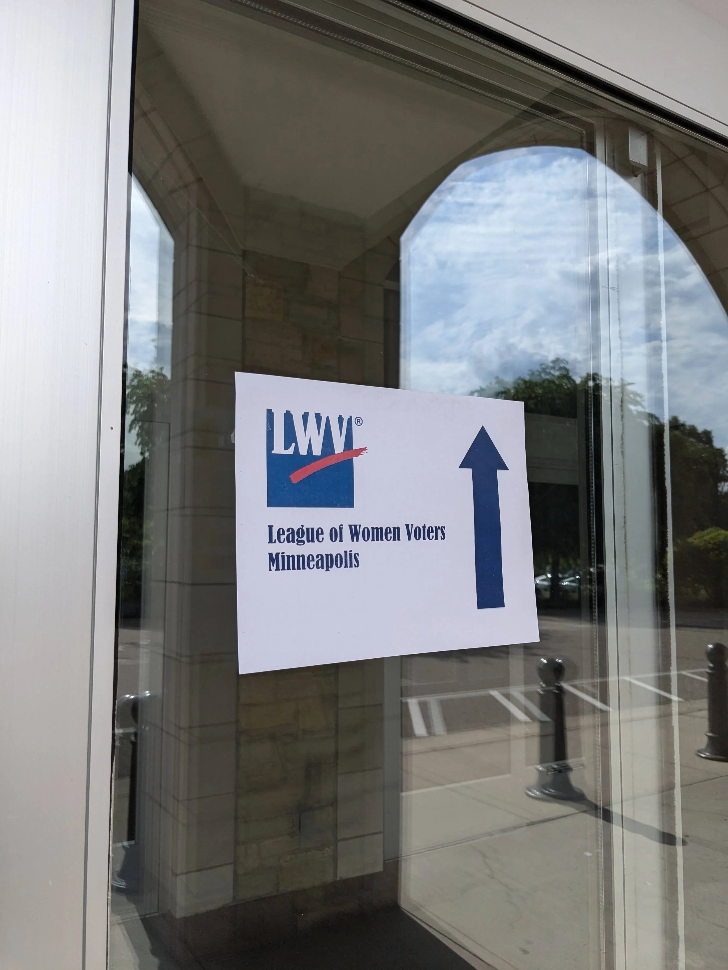Sign on a glass door indicating the League of Women Voters Minneapolis with an arrow pointing upward.