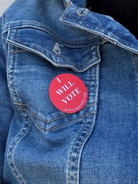 Close-up of a person wearing blue jeans with a red circular sticker that says 'I WILL VOTE' with a website URL.