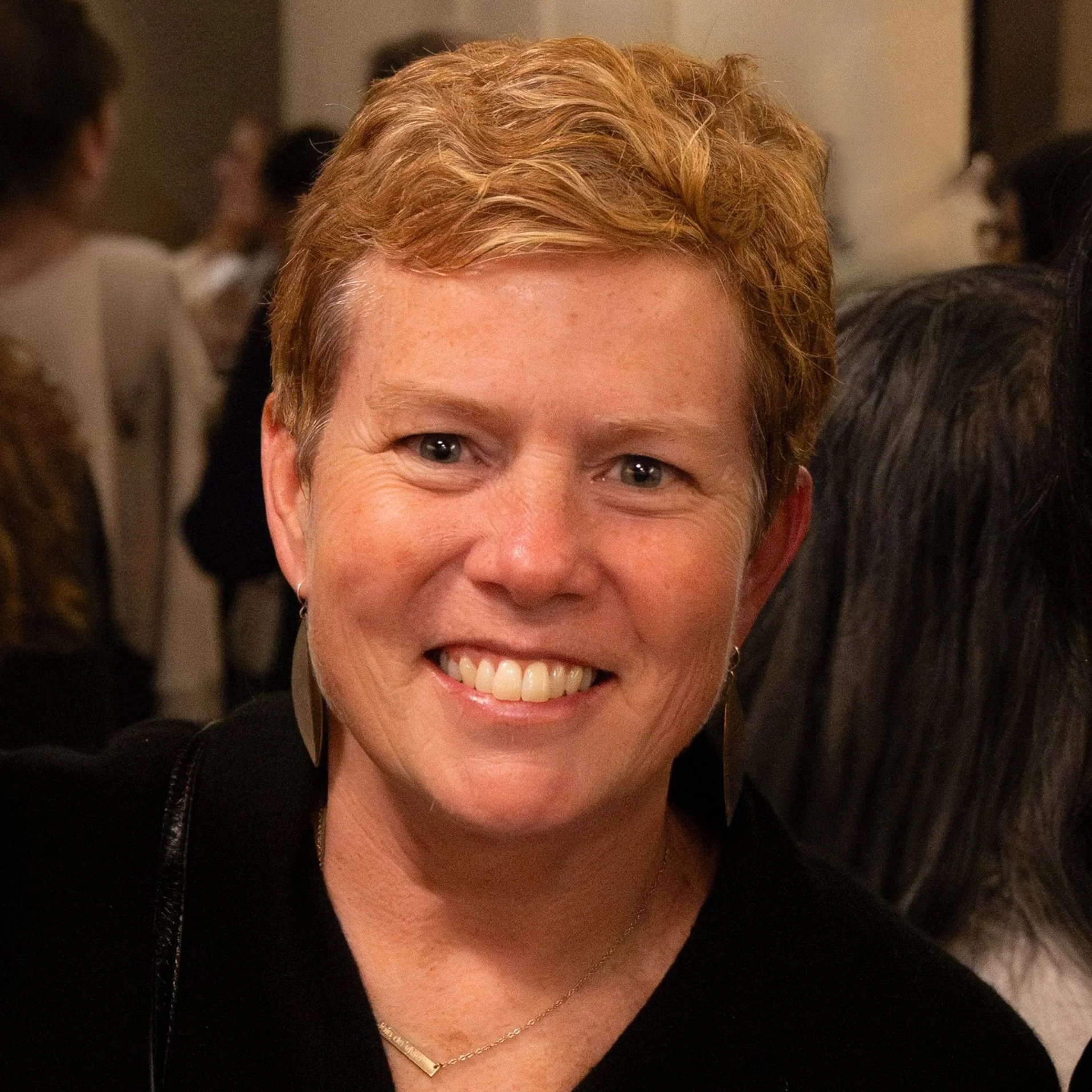 Close-up of a smiling woman with short, curly, reddish hair wearing a black top and a delicate gold necklace.