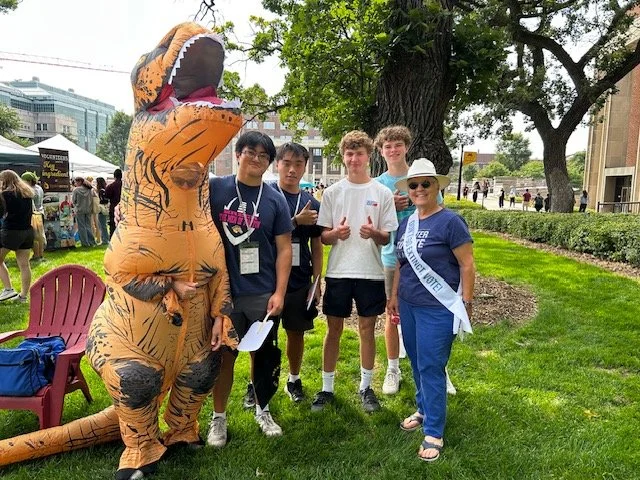 A group of people standing outdoors next to a person in a T-Rex costume at a public event or festival.