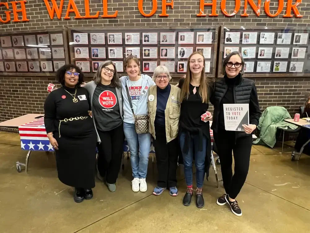 Six women standing side by side in front of a wall featuring multiple framed photographs, with orange letters spelling 'WALL OF HONOR' above. They are smiling, some holding signs or badges, and appear to be at an event encouraging voting.