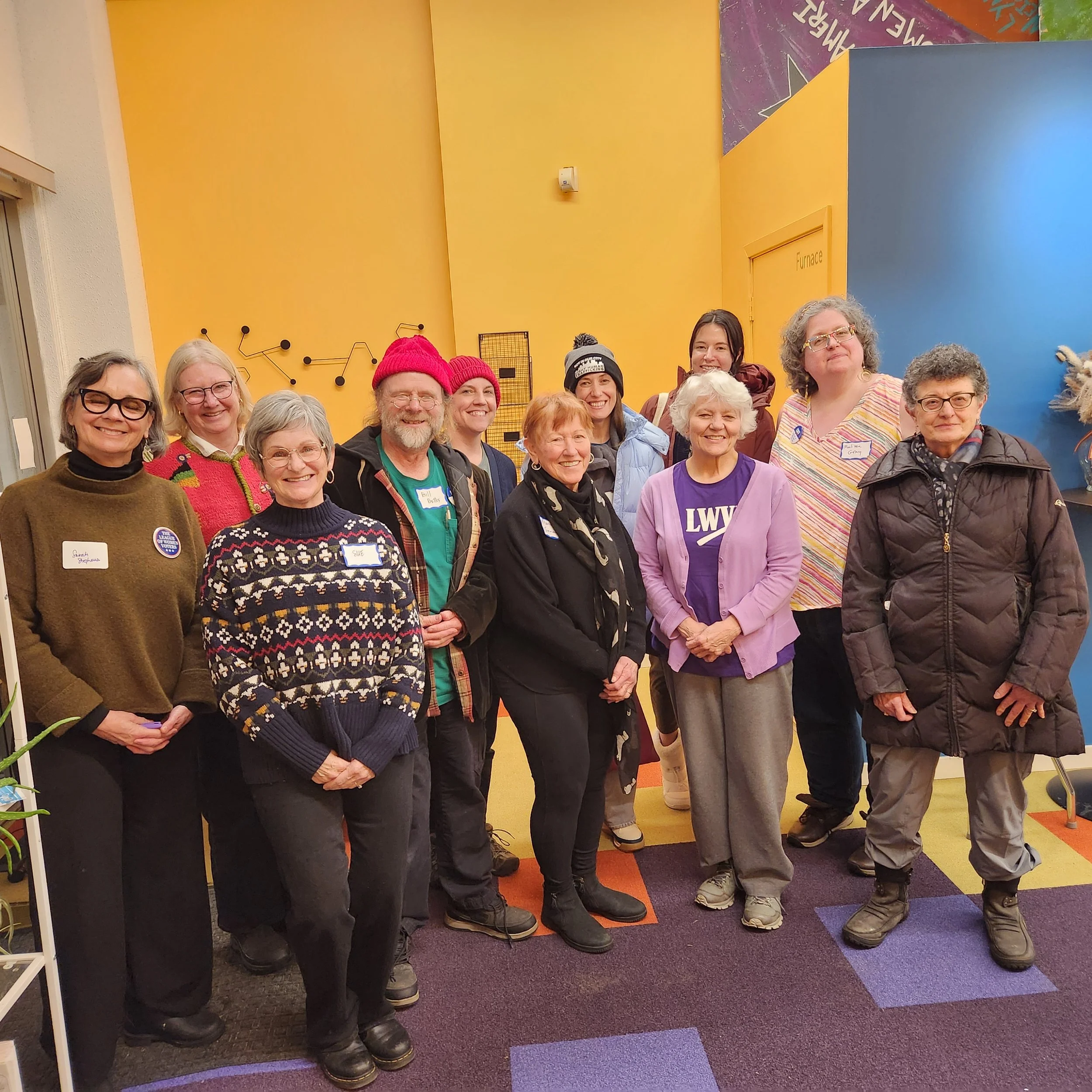 A group of smiling people standing together indoors against a yellow and blue wall, wearing casual clothes and some with name tags, in a colorful room likely at a gathering or event.