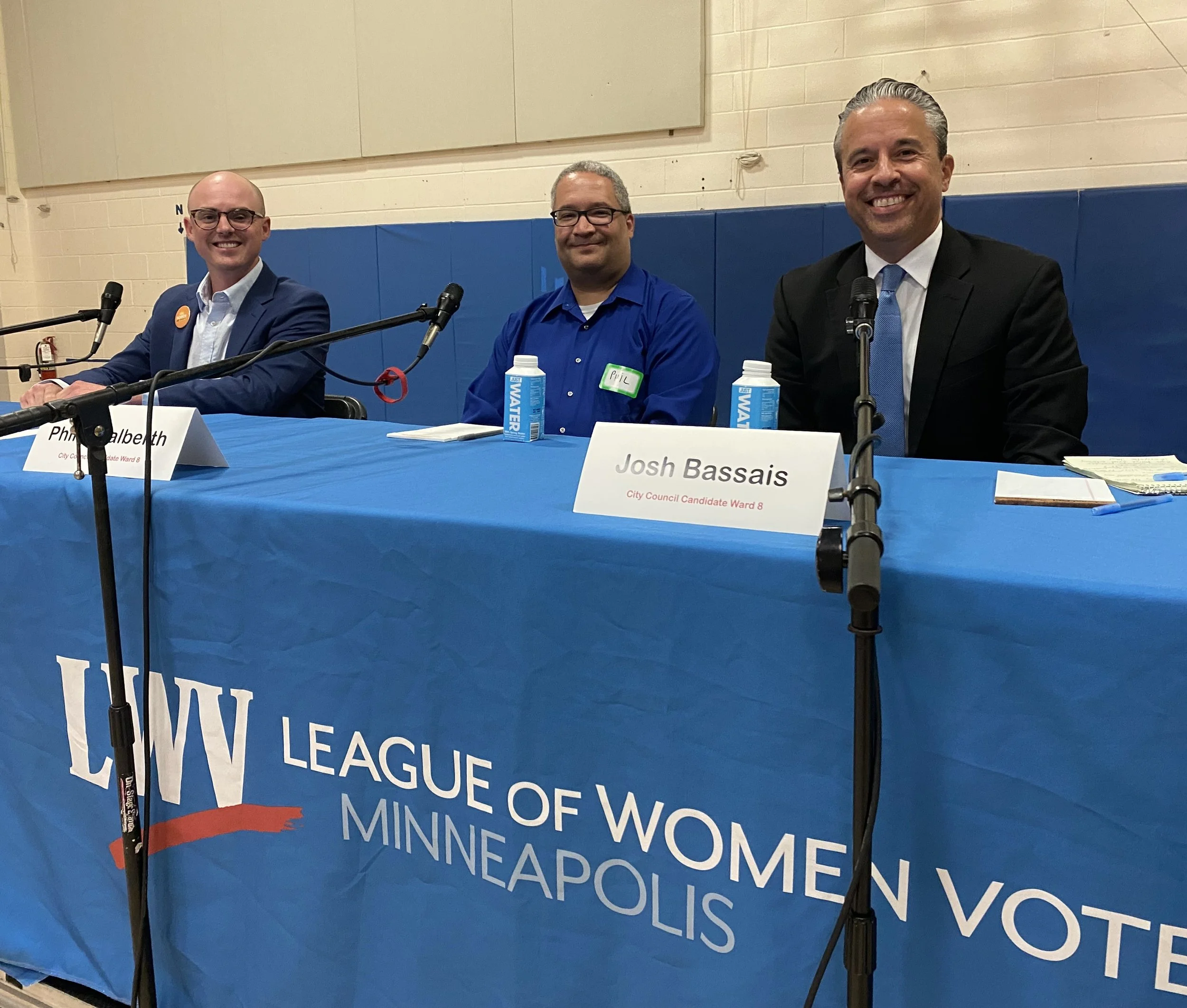 Three men sitting at a table during a political event with a blue cloth displaying the words "League of Women Minnesotans" in the foreground. The man on the left is smiling and wearing glasses, a suit, and a button. The man in the middle is smiling, 