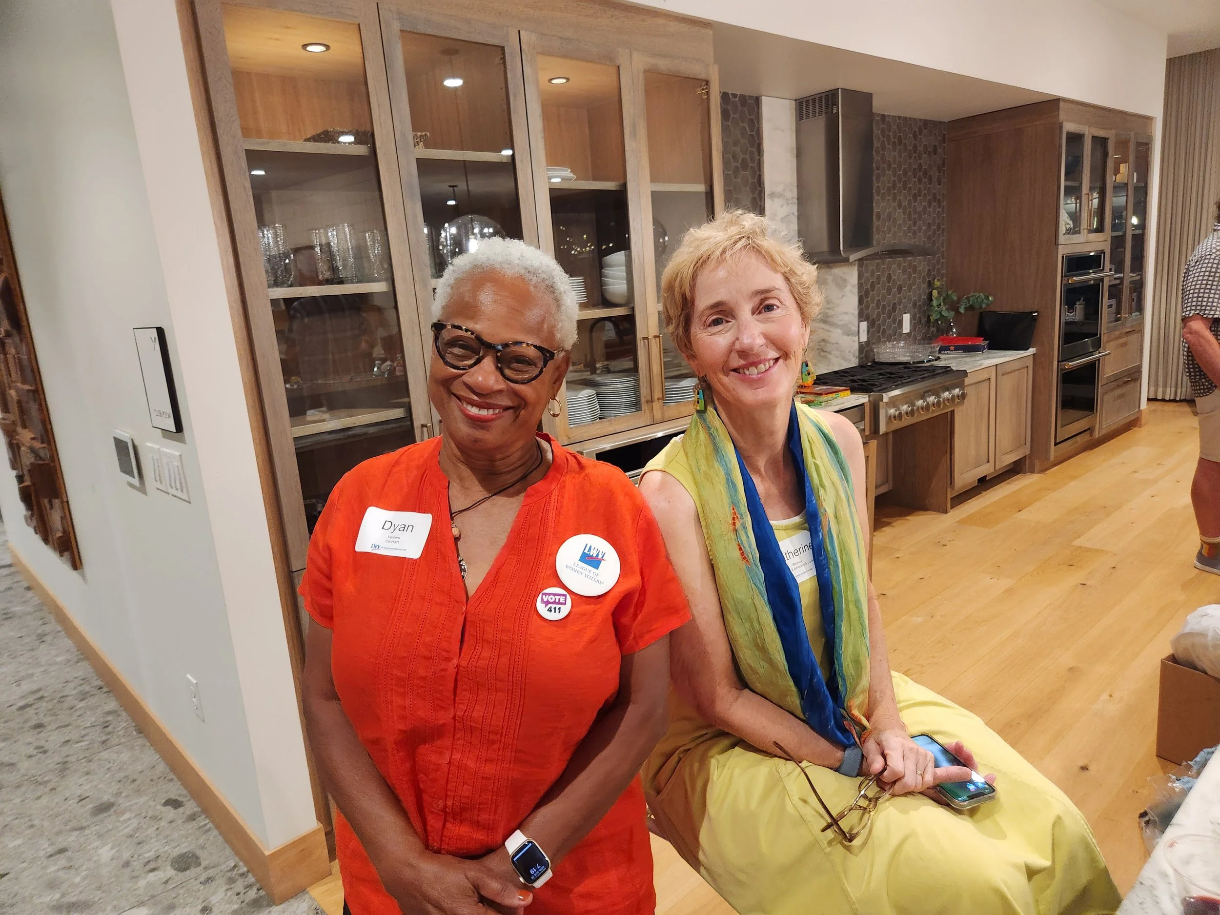 Two smiling women standing in a kitchen, one wearing glasses and an orange top with a name tag that reads "Dyan," and the other with short blond hair wearing a yellow dress and colorful scarf, sitting on a countertop with a smartphone in her hand. Be