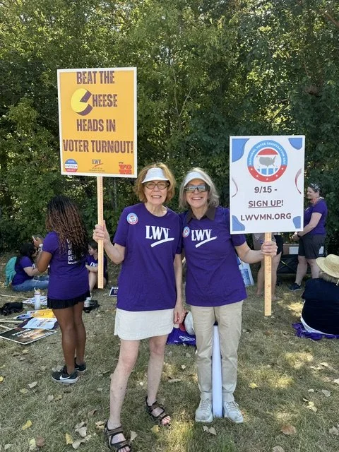 Two women are standing outdoors holding campaign signs for voter turnout and election participation. They are wearing matching purple shirts with 'LWV' on them and sunglasses. Other people are seen in the background, also participating in the event.