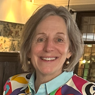 A smiling middle-aged woman with shoulder-length gray hair, wearing a colorful blouse and pearl earrings, inside a warmly lit room with decorative elements in the background.