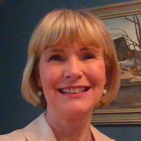 A smiling woman with short blonde hair wearing white earrings and a light-colored blazer, standing in front of a painting with a tree and house.