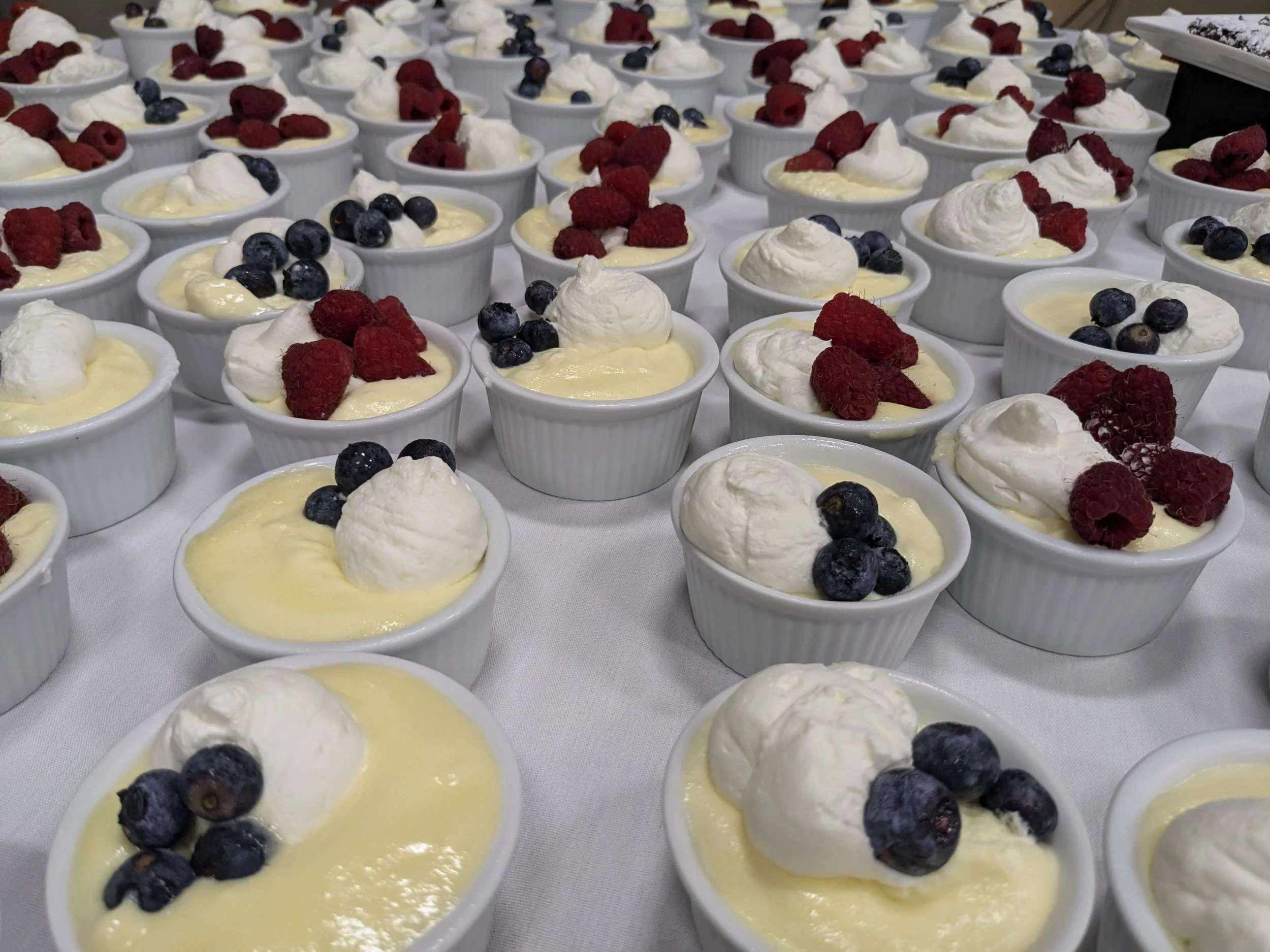 Small white ramekins filled with a yellow pudding, topped with whipped cream and fresh blueberries and raspberries, arranged on a table.